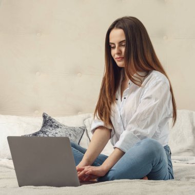 Beyaz gömlek ve kot pantolon giyen ve yatağında otururken laptop kullanan bir kız.