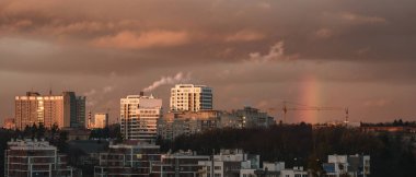 Şehir merkezinde günbatımında gökkuşağı modern binalar, tüten bacalar ve inşaat vinçleriyle