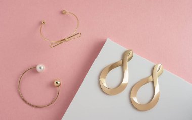 Modern altın bilezikler ve küpeler düz pembe ve beyaz köşeli kağıt arka planda yatıyordu.