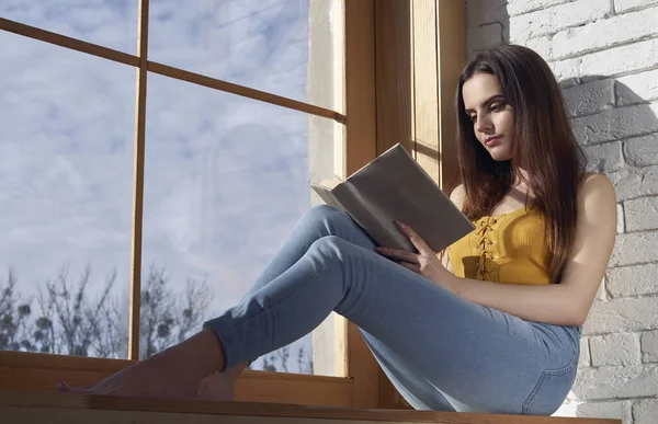 Casual young girl reading a book while sitting on window sill