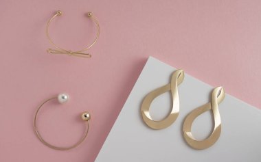 Pembe ve beyaz arka planda iki altın bilezik ve modern küpelerin üst görüntüsü