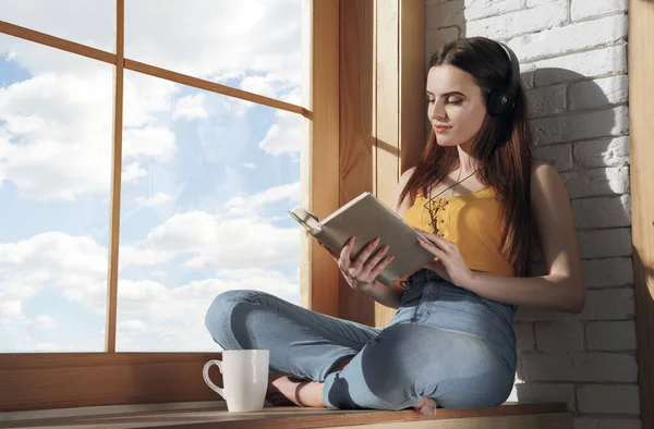 Young casual brunette girl sitting on window sill listening to music and reading a book during quarantine — Stock Photo, Image