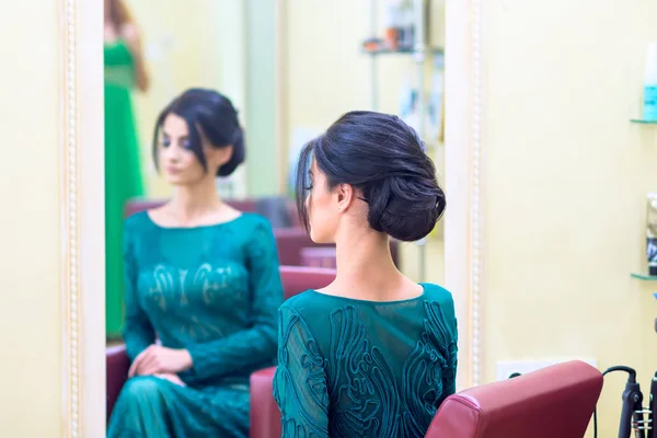 The girl with dark hair and a long dress sitting in the turquoise color barbers chair in front of the mirror after she did my hair. Focus on foreground girl.