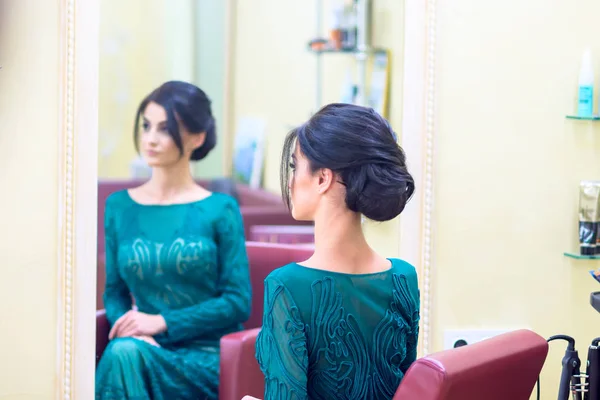 The girl with dark hair and a long dress sitting in the turquoise color barbers chair in front of the mirror after she did my hair. Focus on foreground girl.