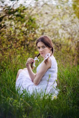 Basit bir genç kız at kuyruklu ve beyaz bir elbise bahar orman çim üzerinde oturuyor.