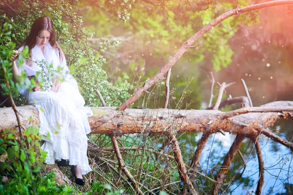 Fiatal lány a fehér köpeny ül egy erdőben egy törött fa naplót. Utómunka. — Stock Fotó