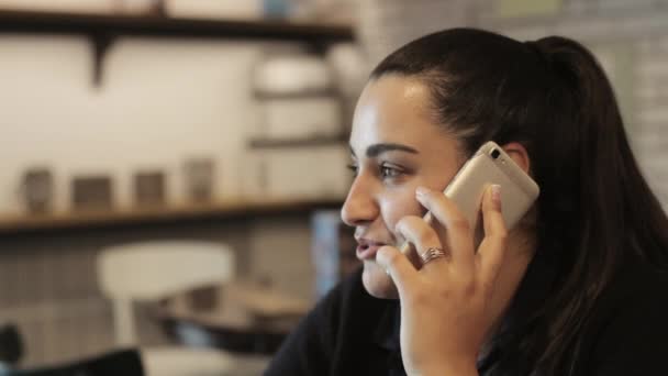 Молодая женщина сидит за столом в кафе и разговаривает по мобильному телефону. — стоковое видео