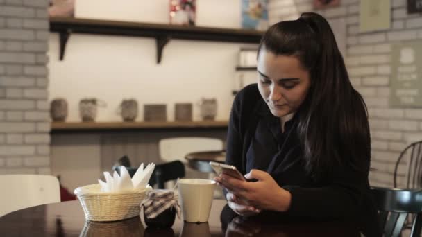 La joven está sentada a la mesa en la cafetería, usando el teléfono inteligente . — Vídeo de stock