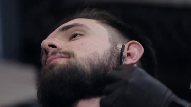 Närbild av en frisör som använder rakapparat för skära skägget av en manlig klient. — Stockvideo