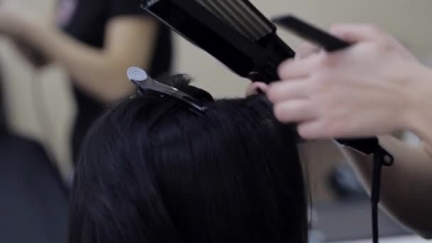 Primer plano de un peluquero alisando el cabello negro con planchas para el cabello . — Vídeo de stock