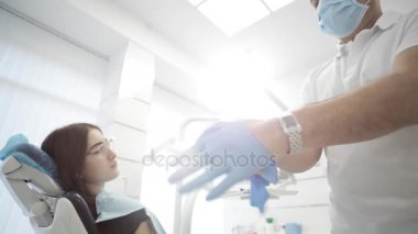 Erkek diş hekimi hastayla arka planda eldivenlerini üzerinde koyarak