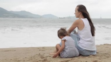 Sırtı kameraya ile kumlu sahilde oturan kızı olan anne.