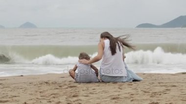 Sırtı kameraya ile kumlu sahilde oturan kızı olan anne.
