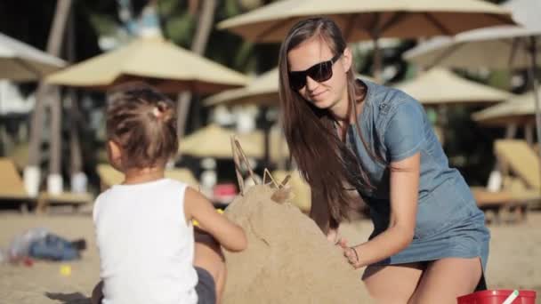 Joven madre en vestido e hija pequeña construyen castillo con arena en la playa . — Vídeos de Stock