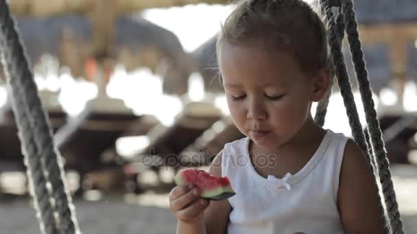 Pequeña niña linda sentada en el columpio y comer sandía roja en la playa de arena — Vídeo de stock