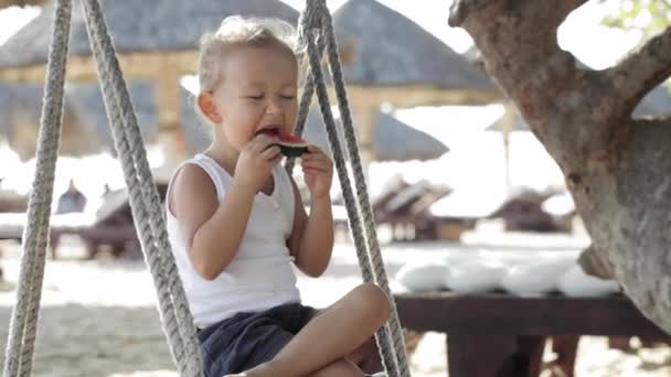 Pequeña niña linda sentada en el columpio y comer sandía roja en la playa de arena — Vídeo de stock