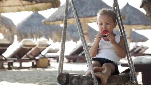 Söta lilla baby flicka sitter på gungan och äta röd vattenmelon på sandstranden — Stockvideo