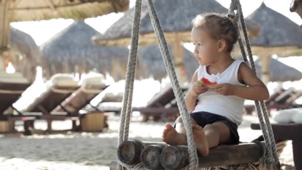 Pequeña niña linda sentada en el columpio y comer sandía roja en la playa de arena — Vídeo de stock