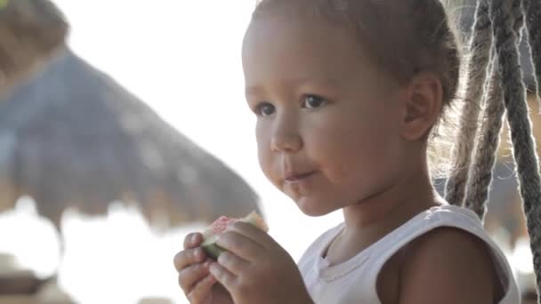 Маленька мила дівчинка сидить на гойдалках і їсть червоний кавун на піщаному пляжі — стокове відео