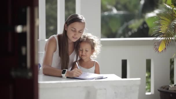 Молодая мама с дочерью рисует на балконе с пальмами на заднем плане — стоковое видео