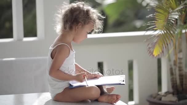 Девушка рисует карандашом на бумаге на балконе с пальмами на заднем плане — стоковое видео