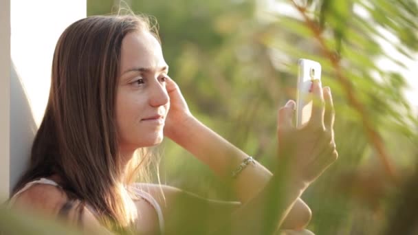 Kobieta za pomocą smartfonu, siedząc na tarasie z ogrodem na tle — Wideo stockowe