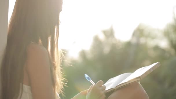 Woman sitting at balcony and writing in diary with garden on the background — Stock Video