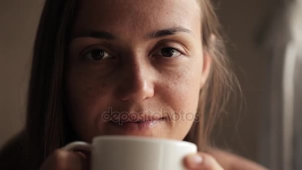 Junge Frau sitzt am Fenster, trinkt Tee und blickt in die Kamera — Stockvideo