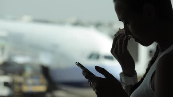 Jonge vrouw met behulp van slimme telefoon en het eten van snacks bij de luchthaven — Stockvideo