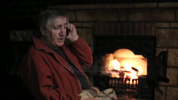Старший говорит по мобильному телефону сидя в кресле перед камином — стоковое видео