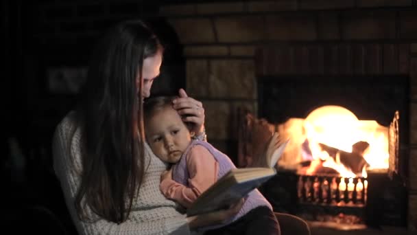 Donna con figlia in grembo seduta davanti al camino a leggere un libro — Video Stock