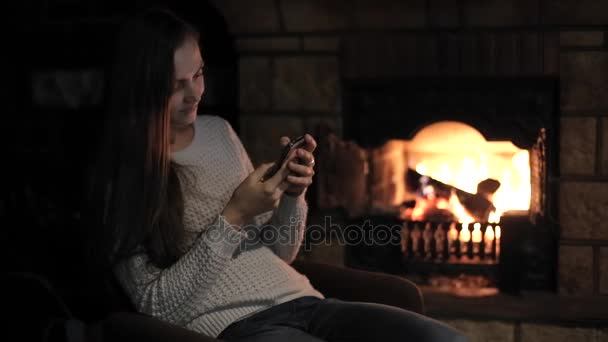 Jeune femme assise avec téléphone intelligent avec la cheminée sur le fond — Video