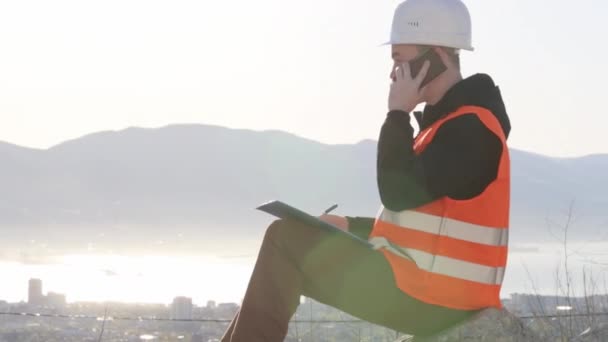 Bouwer praten door slimme telefoon op buiten witing werkplek zitten tegen de zon — Stockvideo