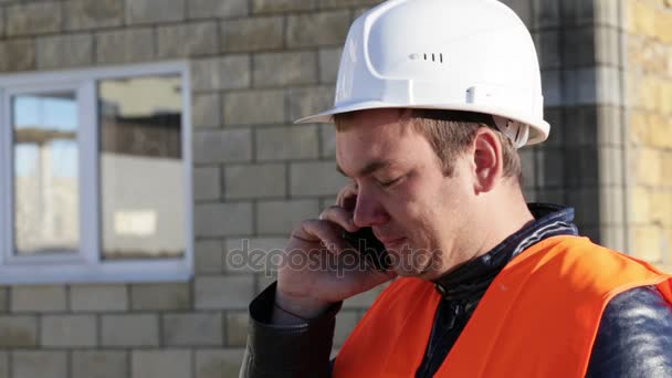 Kavkazská muž v helmu a vestu s úsměvem mluvit po telefonu na pracovišti venkovní — Stock video