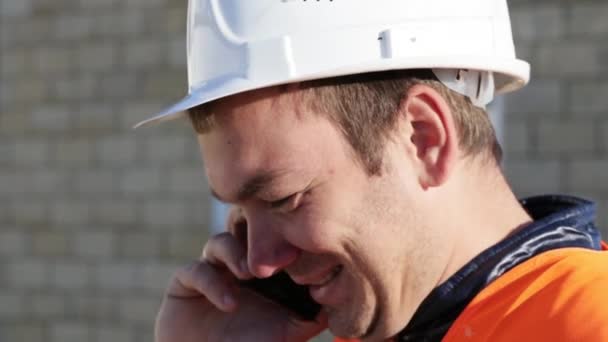 Kavkazská muž v helmu a vestu s úsměvem mluvit po telefonu na pracovišti venkovní — Stock video