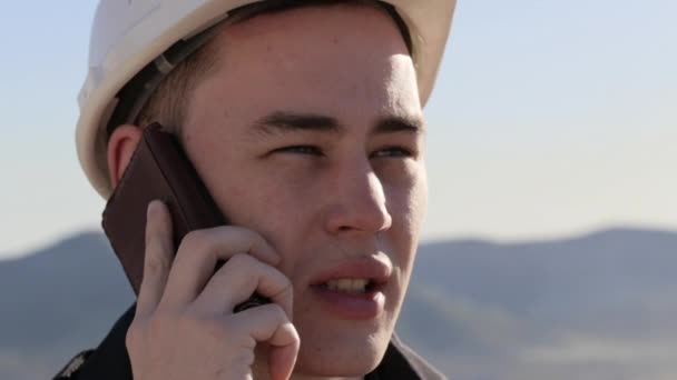 Caucasico uomo in casco e gilet sorridente parlando per telefono sul posto di lavoro all'aperto — Video Stock