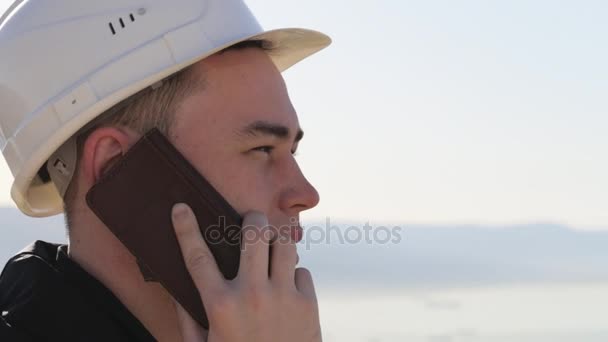 Trabajador caucásico en casco y chaleco sonriendo hablando por teléfono al aire libre — Vídeos de Stock