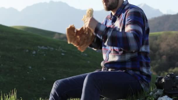 Um homem careca e bonito na natureza come um pão na bela paisagem montanhosa . — Vídeo de Stock