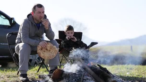 Família de pai e filho comendo pão assado ao lado da fogueira — Vídeo de Stock