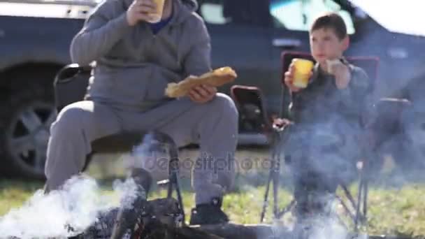 Pai e filho comendo pão torrado e beber refrigerante ao lado da fogueira — Vídeo de Stock