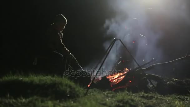 Dos chicos sentados en la noche junto a la hoguera, corrigiendo carbón y hablando . — Vídeo de stock