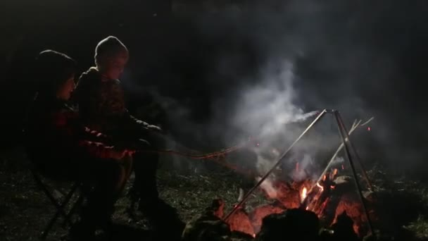 Dos chicos sentados en la noche junto a la hoguera, corrigiendo carbón y hablando . — Vídeos de Stock