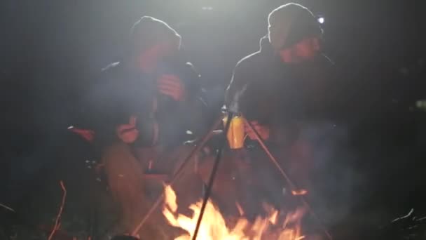 Two friends sits next to bonfire in wood at night, talking and drinking tea. — Stock Video