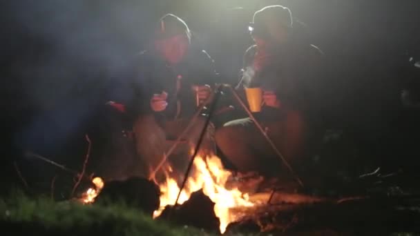 Dois amigos sentam-se ao lado da fogueira em madeira à noite, conversando e bebendo chá . — Vídeo de Stock