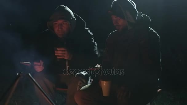 Dos amigos se sientan junto a la hoguera en la madera por la noche, hablando y bebiendo té . — Vídeos de Stock