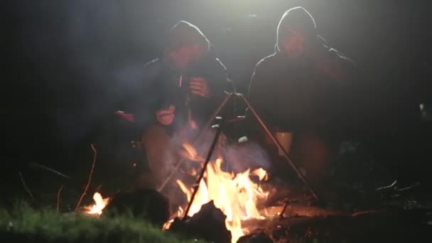 Dos amigos se sientan junto a la hoguera en la madera por la noche, hablando y bebiendo té . — Vídeo de stock