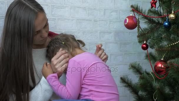 Ung mamma kramar hennes gråtande lilla dotter bredvid granen — Stockvideo