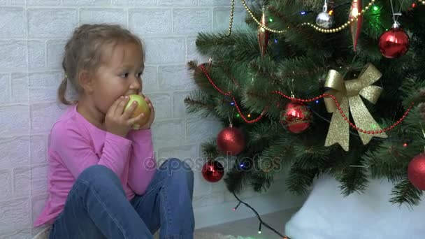 Ładna dziewczynka siedzi na podłodze i jedzenia apple obok choinki. — Wideo stockowe