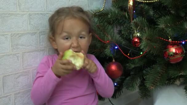 Χαριτωμένο μικρό κορίτσι που κάθεται στο πάτωμα και διατροφικές apple δίπλα στο χριστουγεννιάτικο δέντρο. — Αρχείο Βίντεο