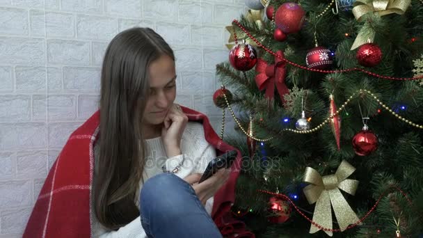 Mujer se sienta en un piso con cuadros rojos y el uso de teléfono inteligente al lado del árbol de Navidad — Vídeos de Stock